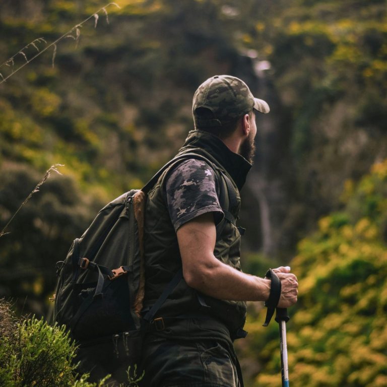 Man in camouflage stands on a trail, looking at a waterfall surrounded by greenery.